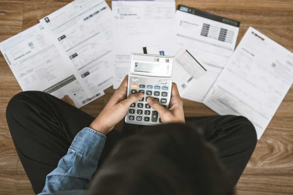 A person sits on the floor in front of paperwork holding a calculator.