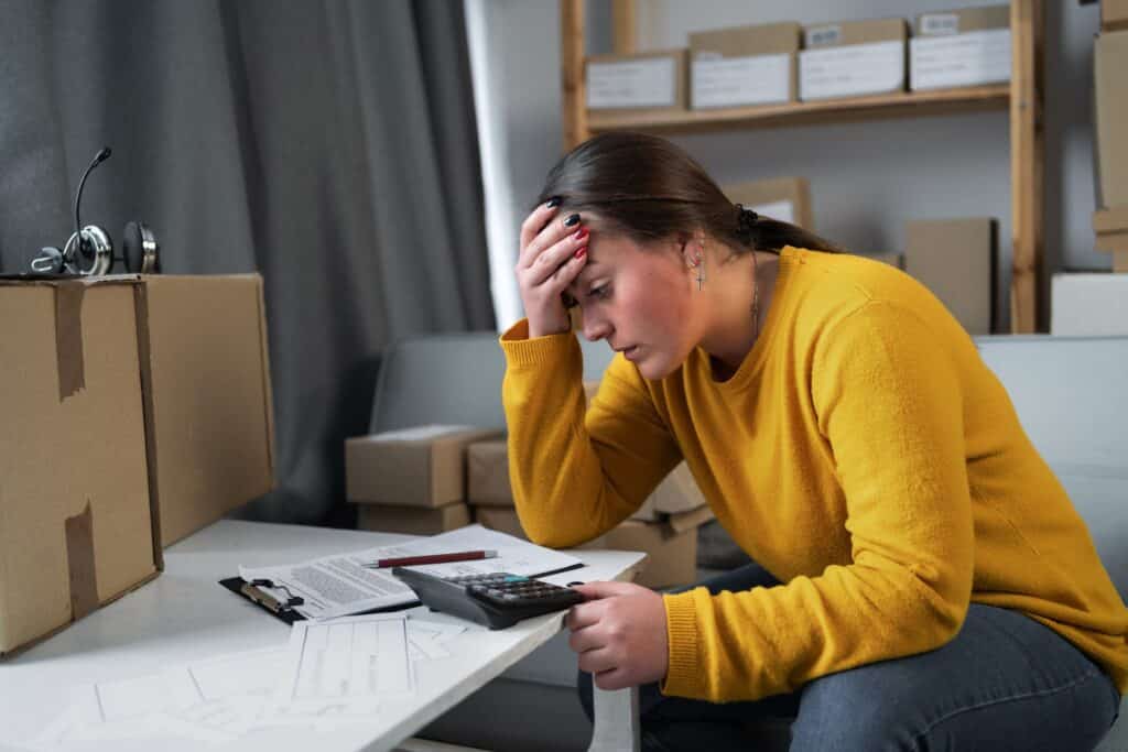 A woman in a yellow blouse sitting down with her hand on her head looking at a calculator and documents