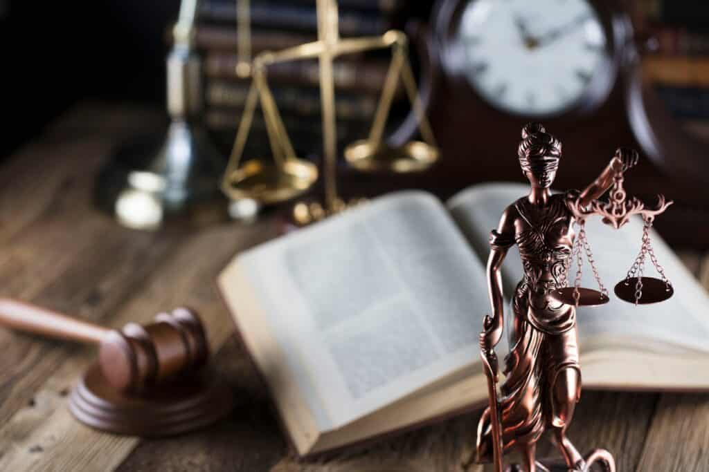 A statue of lady justice sits next to an open law book, a gavel resting on its stand, and scales of justice.