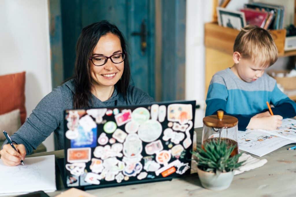 Child at a desk writing in a notebook while woman next to him is on a laptop