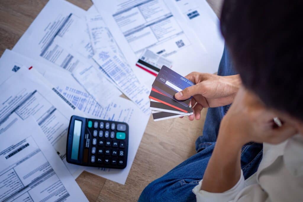 Person sitting on the floor holding credit cards in his right hand and sitting in front of bills and a calculator