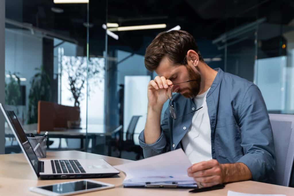 Man sitting at a desk with his glasses in his hand and his hand on his head. Sitting in front of a computer and a clipboard of paperwork