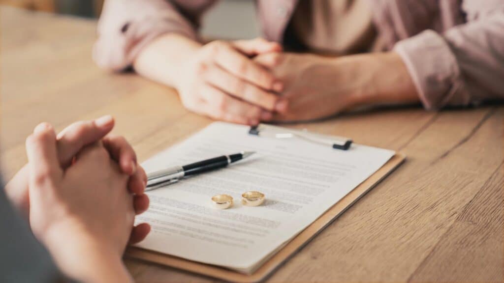 Two people sit across from each other at a wooden table in front of a clipboard with two wedding rings and a pen resting on it.